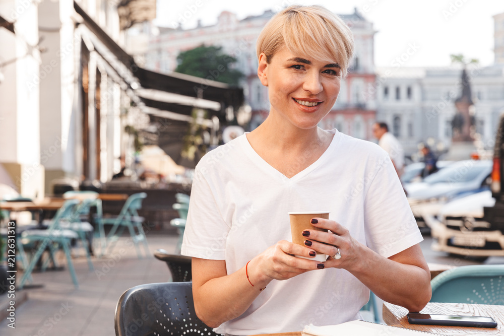 Image of cheerful blonde woman wearing white t-shirt sitting in street cafe in summer, and drinking coffee from paper cup