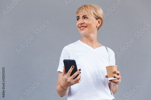 Image of cheerful blond woman wearing casual clothing smiling while holding coffee from paper cup and smartphone, isolated over gray wall