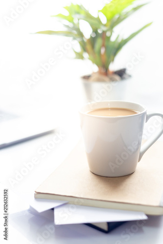 Coffee cup on notebook with laptop on white table in the morning sun light  Working space