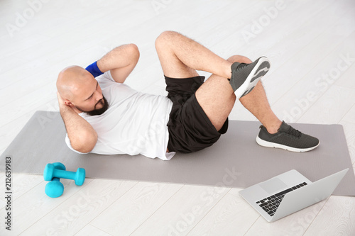 Overweight man doing exercise while watching tutorial on laptop at home