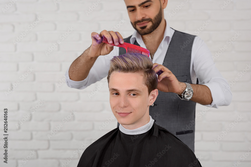 Hairdresser doing hair violet colored toning for young boy. Barber using  red plastic comb, wearing white shirt and grey waistcoat, having violet  color spots on both hands. White brick background. Stock Photo |