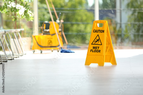 Safety sign with phrase Caution wet floor and blurred mop bucket on background. Cleaning service photo