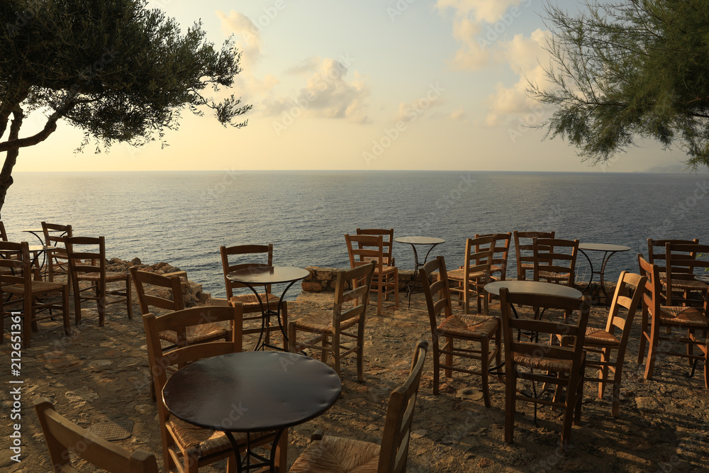 Iron tables and wooden chairs, typical traditional greek cafes, overlooking the roofs of houses and the sea in Monemvasia, Peloponnese, Greece, June 2018.