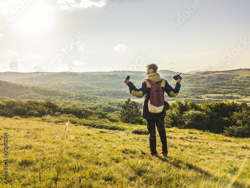 portrait of young man with photography equipment and raised hands walking on the mountain field © Mihail