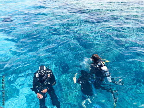 Three underwater divers in black scuba diving suits, a man and a woman with oxygen bottles sink under the transparent blue water in the sea, the ocean in a tropical paradise warm resort.