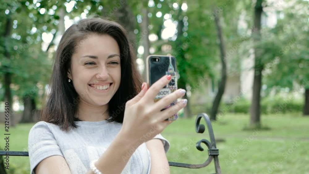 The girl corrects hairstyle by looking at the smartphone as mirror, in the park The girl corrects hairstyle by looking at the smartphone as mirror, in the park