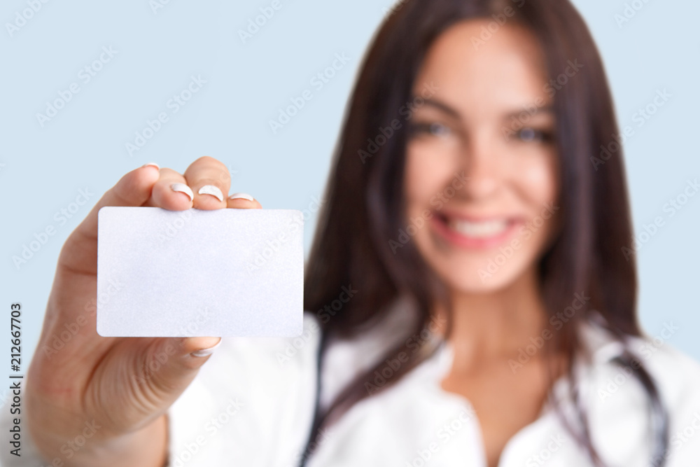 Horizontal shot of medical worker holds blank card, blurred silhouette, free space for your name and surname. Cute female nurse holds name tag. Professional young woman therapist stands indoor Horizontal shot of medical worker holds blank card, blurred silhouette, free space for your name and surname. Cute female nurse holds name tag. Professional young woman therapist stands indoor