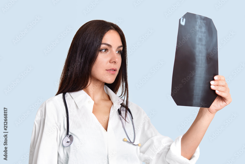 Serious female doctor looks attentively at X ray, examines people`s spine, wears white lab gown, has phonendosope on neck, stands sideways against light blue background. Medical treatment concept Serious female doctor looks attentively at X ray, examines people`s spine, wears white lab gown, has phonendosope on neck, stands sideways against light blue background. Medical treatment concept