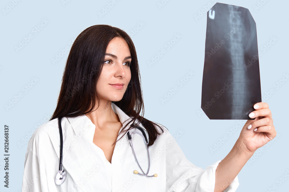 Indoor shotof pleasant looking female nurse or doctor looks attentively and happily at X ray, studies skeleton, dressed in white medical robe. Professional radiologist in hospital. Health care concept Indoor shotof pleasant looking female nurse or doctor looks attentively and happily at X ray, studies skeleton, dressed in white medical robe. Professional radiologist in hospital. Health care concept