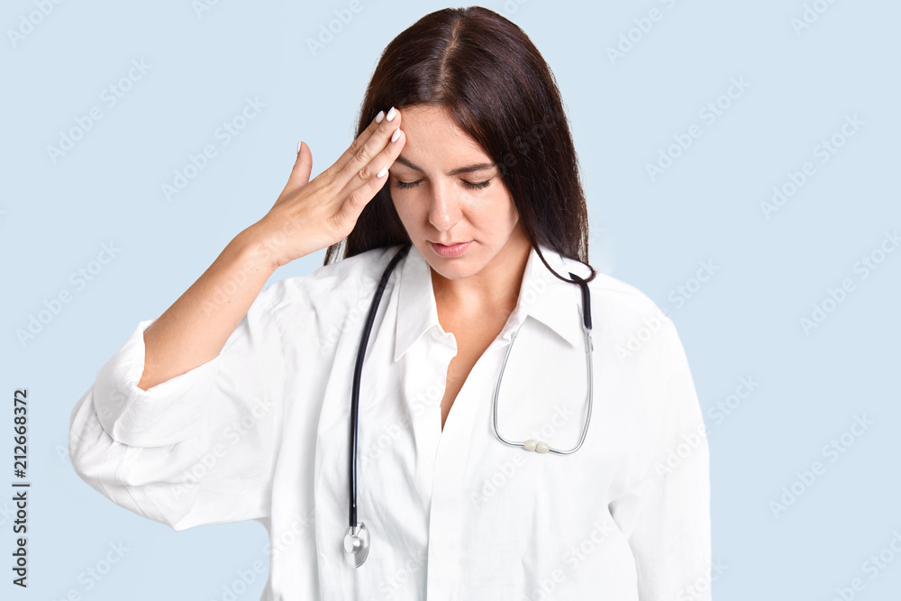 Attractive brunette young female nurse has headache after making diagnosis, keeps hand on forehead, concentrated down, dressed in white robe, isolated over blue background. Overworked doctor Attractive brunette young female nurse has headache after making diagnosis, keeps hand on forehead, concentrated down, dressed in white robe, isolated over blue background. Overworked doctor