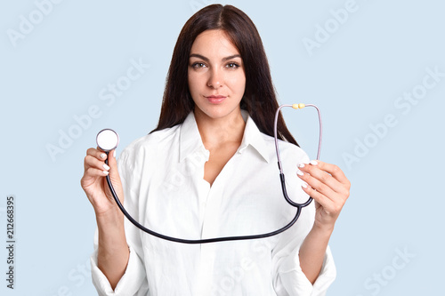 Horizontal shot of beautiful professional female therapist with stethoscope, dressed in white gown, going to examine patient, has serious expression, stands against blue background. Medical treatment photo