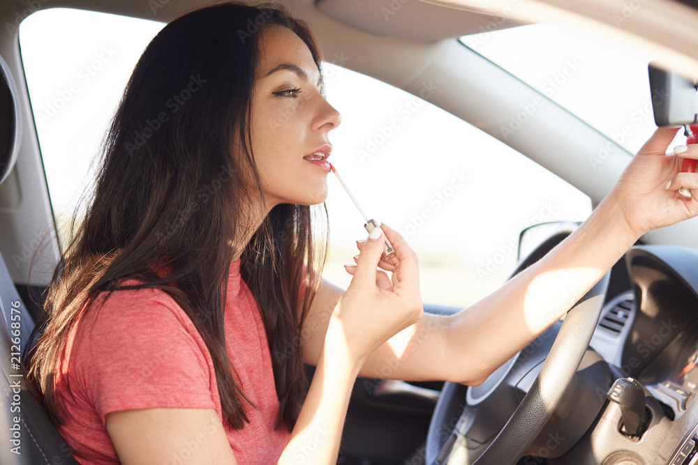 Sideways shot of pretty brunette female applies lipstick, poses in car, has no time for make up, prepares for important meeting, dressed in casual t shirt. People, beauty, transport concept.