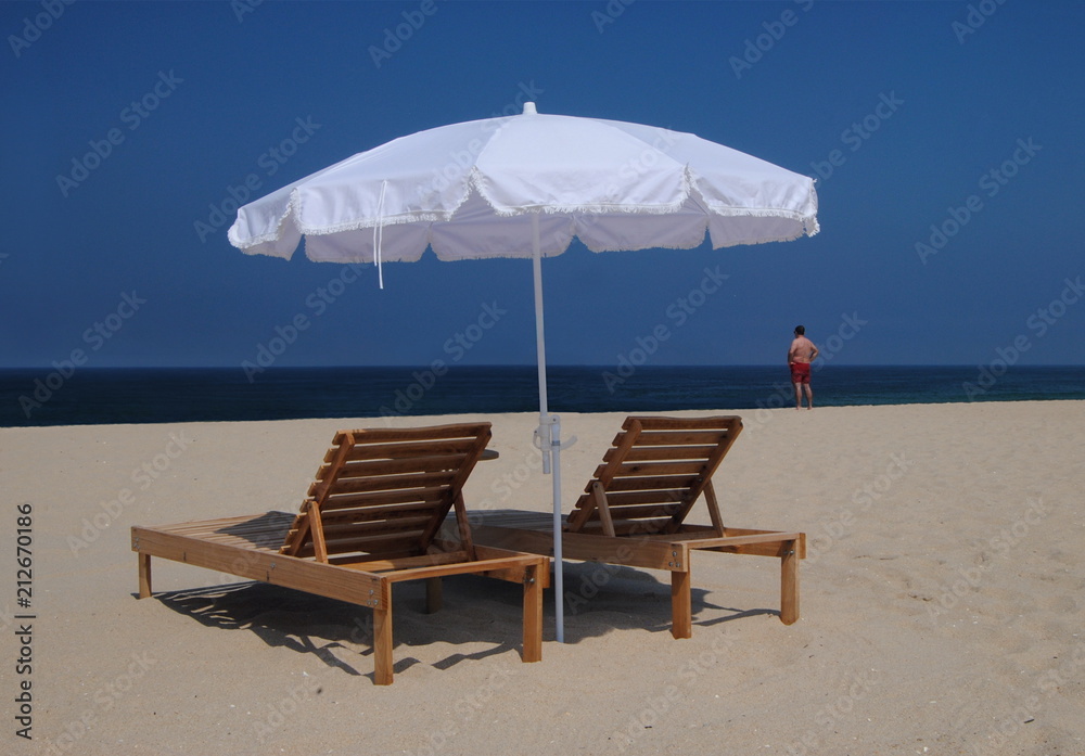 Espreguiçadeiras na praia - com um guarda-sol branco e uma pessoa do sexo masculino ao longe com calções vermelhos