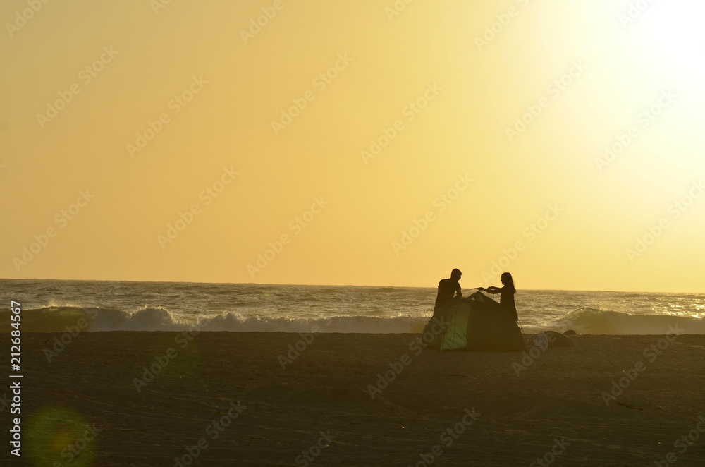 man and woman young couple silhouettes on beach putting up camping tent at dusk