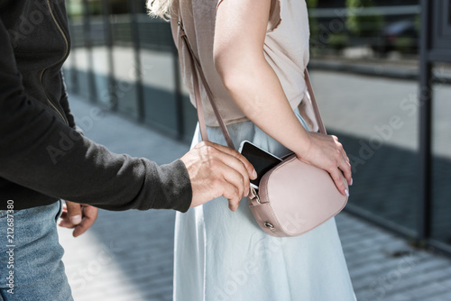 cropped view of criminal man pickpocketing smartphone from womans bag on street