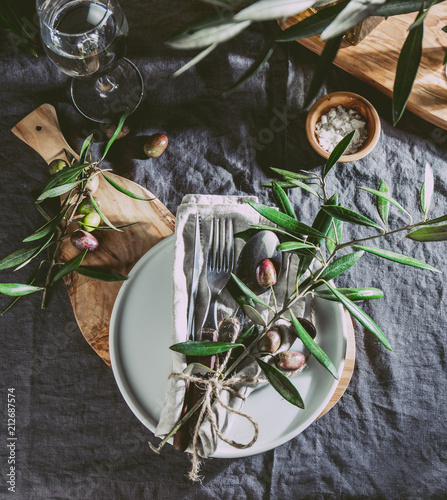 Top view of table setting with gray Linen tablecloth and napkin, white plate, cutlery and and olive tree branch boards decoration. Top view photo