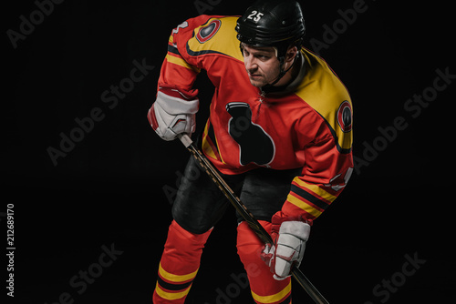 professional hockey player in protective sportswear holding hockey stick and looking away isolated on black