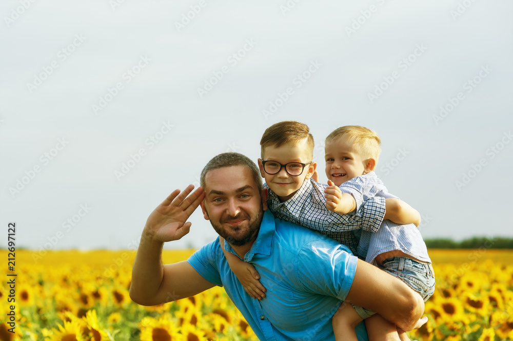 Cheerful father with his sons on vacation in the field with sunflowers . The parent plays with his son . Father's day Cheerful father with his sons on vacation in the field with sunflowers . The parent plays with his son . Father's day