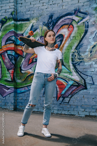 attractive girl with tattoos holding skateboard over shoulder near wall with graffiti