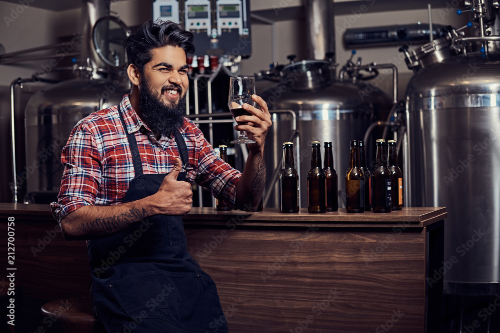 Stylish full bearded Indian man in a fleece shirt and apron holds a glass of beer, sitting behind the counter in the brewery. Stylish full bearded Indian man in a fleece shirt and apron holds a glass of beer, sitting behind the counter in the brewery.
