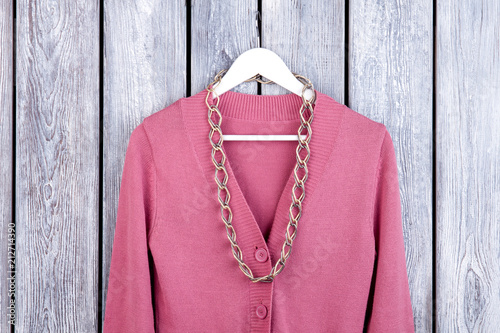Flat lay wool coat with accessorie. Pink cardigan with necklace on hanger. Dark wooden desk surface background.