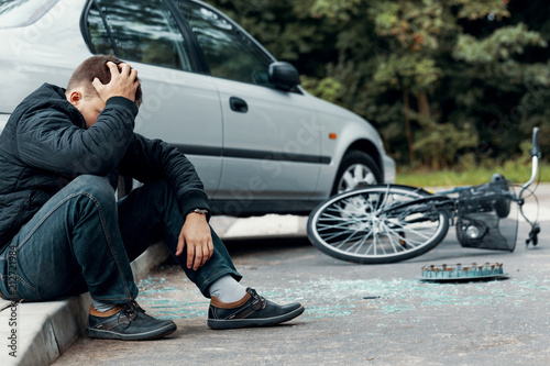 Worried biker holding his head and sitting on a pavement next to a car and bike crash
