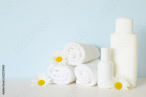 White bottles and towels on white shelf in bathroom. Beautiful camomiles or daisies. Fresh flowers. Care about clean and soft face  hands  legs and body skin. Empty place for text on pastel blue wall.