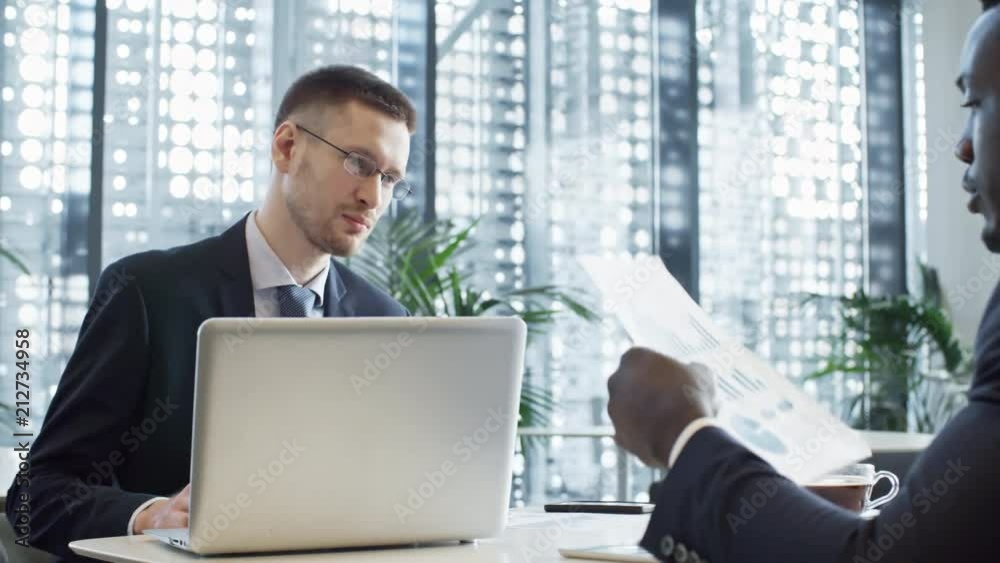 Young Caucasian businessman reading financial documents while working with African American colleague in large office