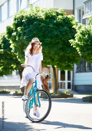 Young female cyclist on the city street on a sunny summer day. A girl in a light dress and sunglasses on a vintage blue bicycle is not hiding admiration © anatoliy_gleb
