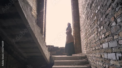 Young refugee muslim woman in hijab standing alone in abandoned building and looking around, worried and terrified photo