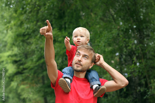 Cute toddler boy sits on his father's shoulders. They point fingers in the same direction. Fatherhood concept. Family look clothing photo