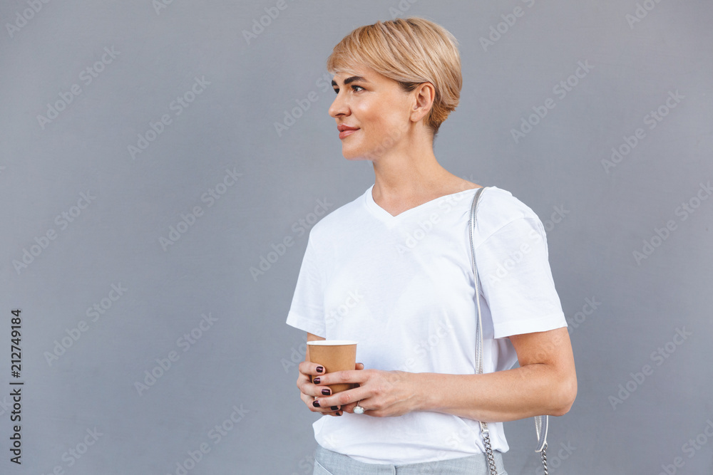 Image closeup of beautiful blond woman wearing white t-shirt standing isolated over gray wall, and holding coffee from paper cup Image closeup of beautiful blond woman wearing white t-shirt standing isolated over gray wall, and holding coffee from paper cup
