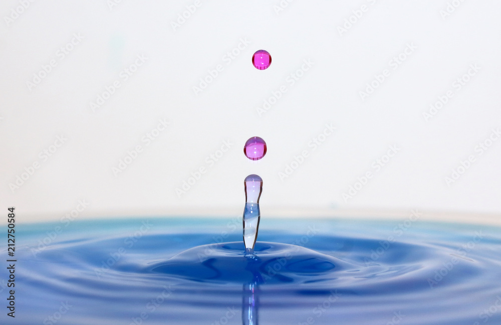 A drop of pink water creates ripples in blue water on a white background. A drop of pink water creates ripples in blue water on a white background.