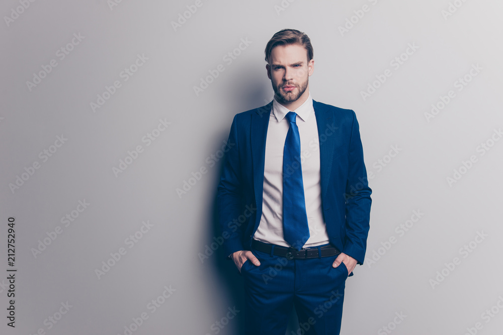 Fototapeta premium Portrait of stunning, attractive, sexy, brutal man in blue suit with tie with stubble, holding two hands in pockets of pants, looking ta camera, isolated on grey background