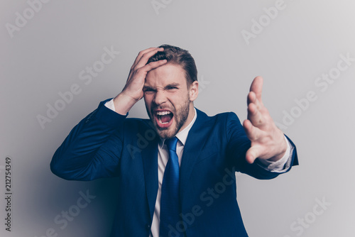 Portrait of violent, stylish, attractive, angry man in blue suit, tie with bristle, hairstyle, gesture palm front, yelling with wide open mouth, holding hand on head, isolated on grey background