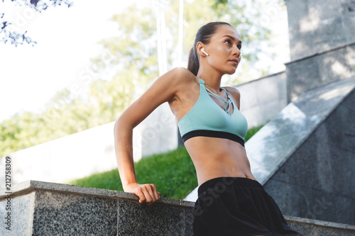 Portrait of beautiful sporty woman 20s in sportswear looking aside, and listening to music with bluetooth earbud while walking in green park photo