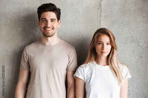 Portrait of young caucasian people man and woman in basic clothing posing together at camera with happy smile, isolated over concrete gray wall indoor