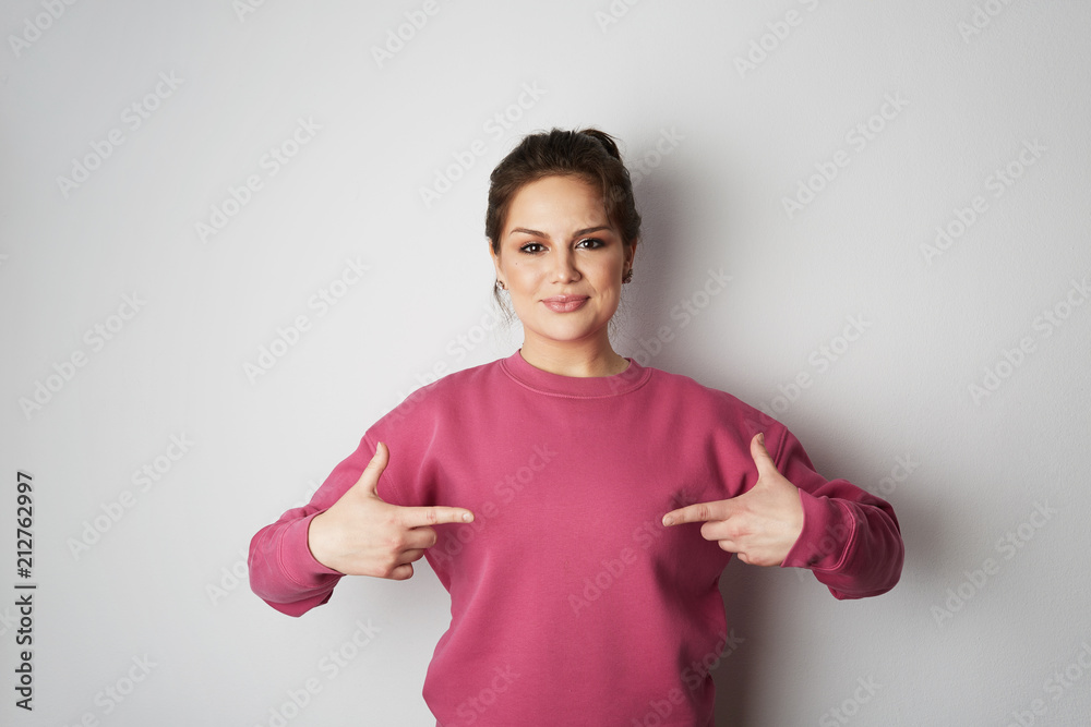 Beautiful smiling hipster girl wearing pink hoody and pointing finger with space for your logo or design. Mock-up of pink hoody on white empty wall in the background. Copy paste text space