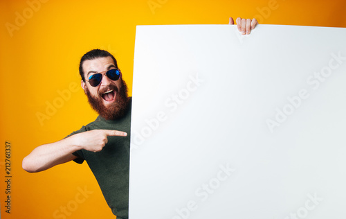Excited bearded man wearing sunglasses is hiding behind and pointing at white blank sheet on yellow background. photo