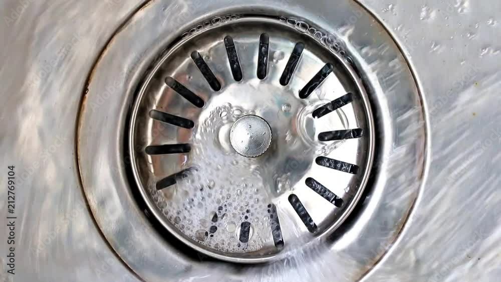 Water with soap bubbles flows into the drain hole in the metal sink, macro view. Mechanically adjustable drain plug closeup. White foam with bubbles of cleaner in a washbasin Water with soap bubbles flows into the drain hole in the metal sink, macro view. Mechanically adjustable drain plug closeup. White foam with bubbles of cleaner in a washbasin
