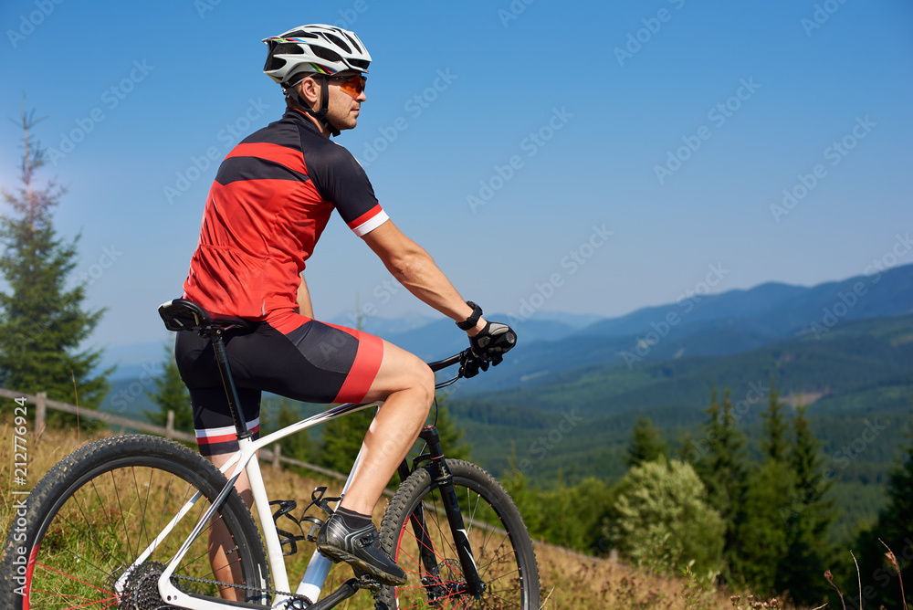 Active professional sportsman cyclist stopped bicycle on top of hill to enjoy beautiful view of distant Carpathian mountains on blue summer sky background. Active lifestyle and extreme sport concept.
