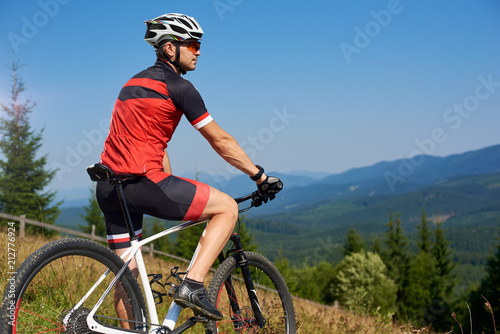 Active professional sportsman cyclist stopped bicycle on top of hill to enjoy beautiful view of distant Carpathian mountains on blue summer sky background. Active lifestyle and extreme sport concept.
