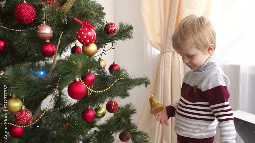 A little boy in a warm sweater stands near a Christmas tree and looks at Christmas toys. Portrait of a little boy near a Christmas tree.
