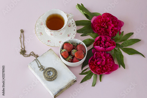 on a pink background, three pink peonies, a cup of tea, a strawberry in a vase, a book and an old pocket watch