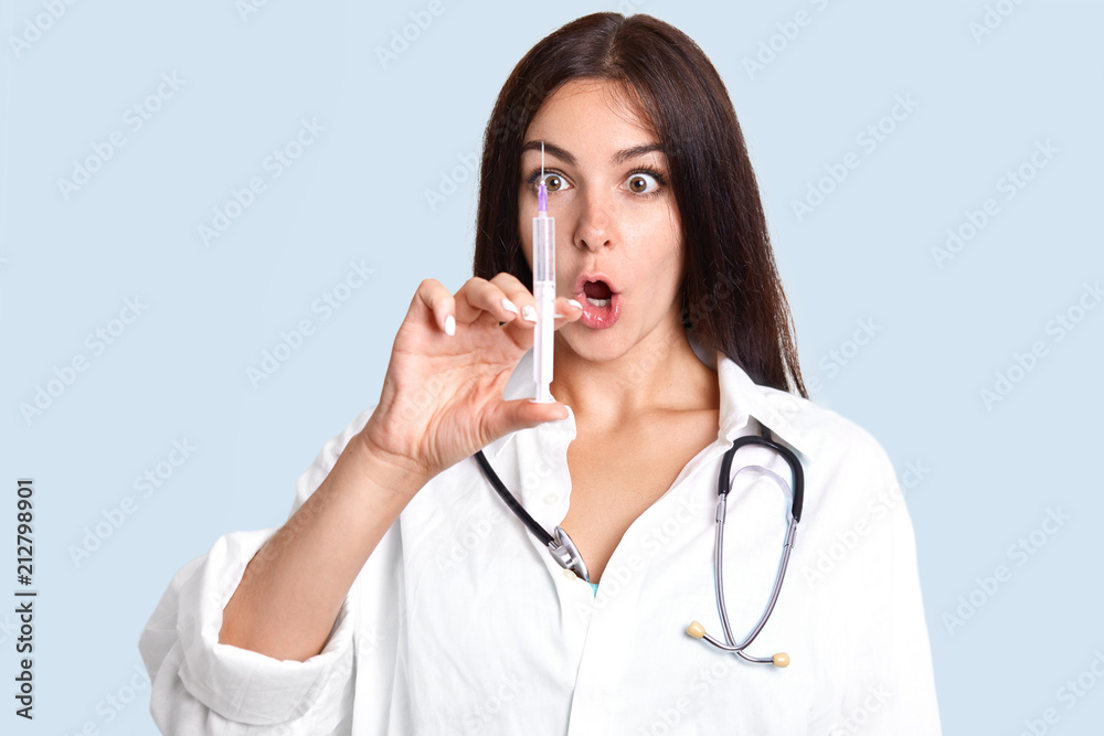 Surprised brunette female nurse looks terrified on syringe with serum, going to make injection for patient, wears white gown and stethoscope, isolated over blue background. Medicine concept.