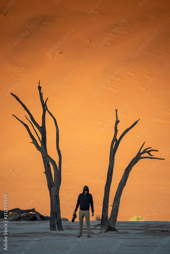 Young man photographer and traveler standing between dead trees in deadvlei (Sossusvlei) during sunrise, famous natural landmark in Namib desert of Namibia, Africa