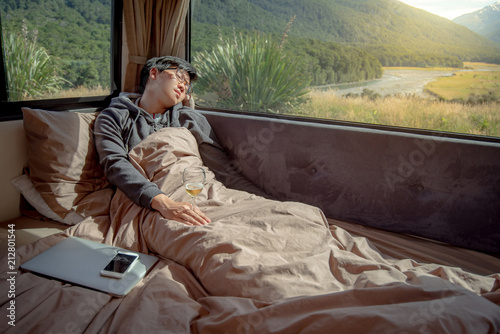 Young Asian man hangover after drinking beer in camper van with mountain scenic view through the window, laptop computer and smartphone on the bed. digital nomad on road trip concept