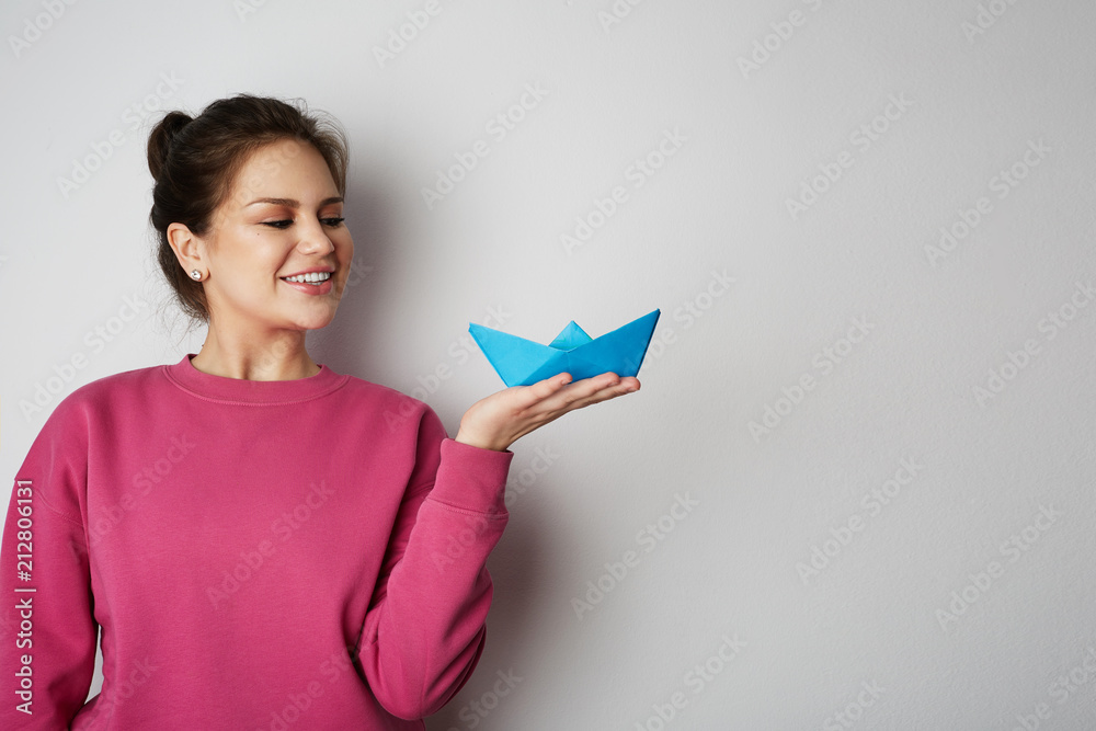 Smiling woman holding a blue color paper ship on empty gray background.Emotions, people, beauty, fashion and lifestyle, travel, tourism and people concept. Copy paste text Smiling woman holding a blue color paper ship on empty gray background.Emotions, people, beauty, fashion and lifestyle, travel, tourism and people concept. Copy paste text