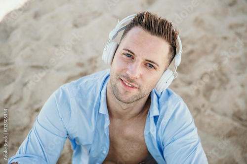 Portrait of Young hipster man in a blue shirt and jeans listening to music in headphones on a smartphone and is sitting on beach.Digital Music lounge and relaxing concept.Closeup