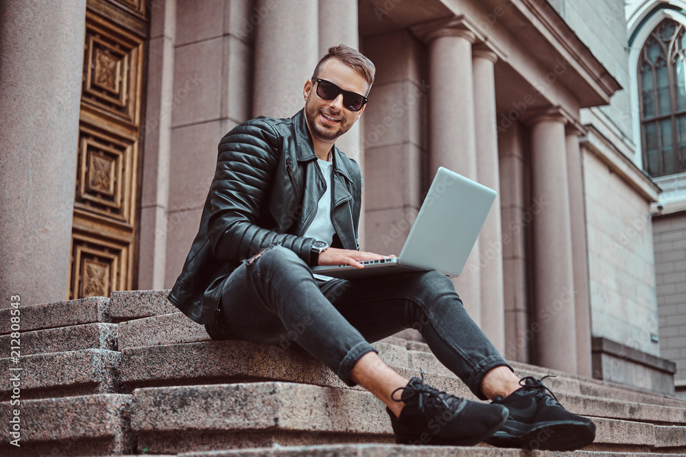 Smiling fashionable freelancer guy dressed in a black jacket and jeans holds a laptop while sitting on steps against an old building in Europe.
